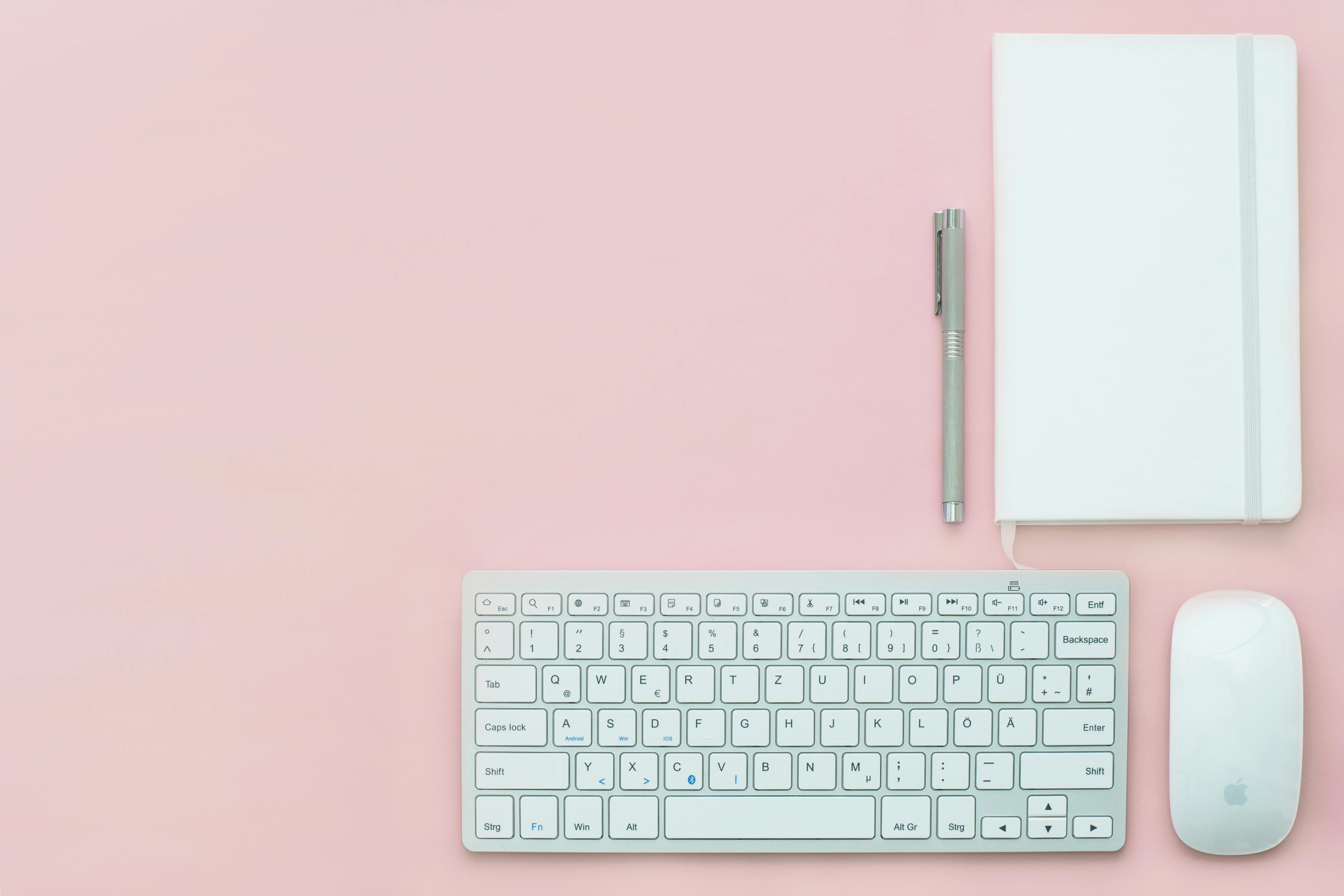 Flat lay of a minimalist workspace featuring a keyboard, notebook, and pen on a pink surface.
